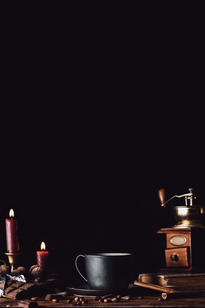 Close-up van kop koffie aan houten tafel met truffels, korrels van koffie, chocolade en kaarsen op zwarte achtergrond  - Foto, afbeelding