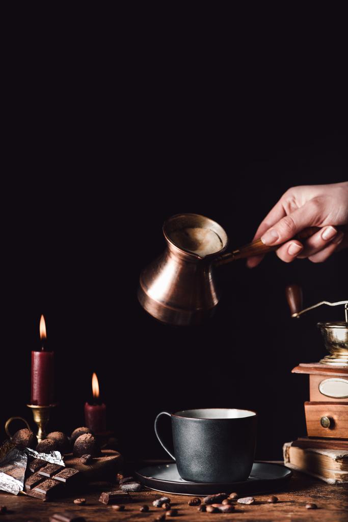 Teilansicht einer Frau, die Kaffee vom Türken in die Tasse am Tisch mit Schokolade, Trüffeln, Kerzen und Kaffeekörnern auf schwarzem Hintergrund gießt - Foto, Bild