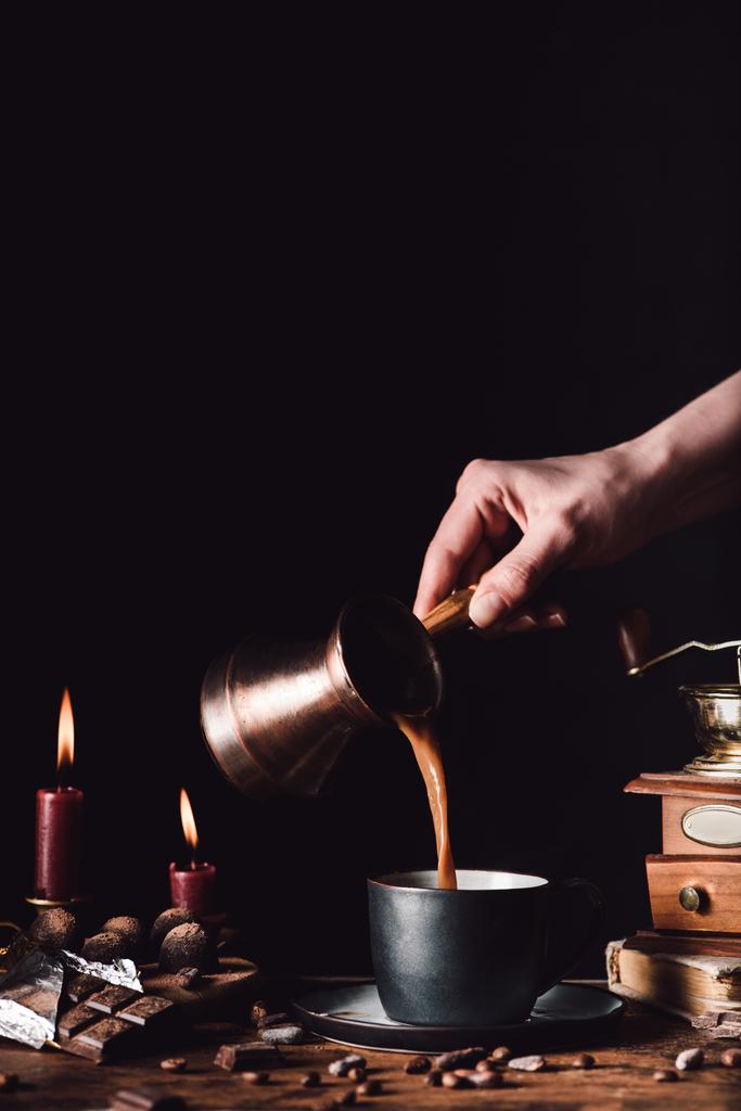 bijgesneden schot vrouw gieten koffie uit turk in beker aan tafel met koffie, chocolade, truffels en kaarsen korrels op zwarte achtergrond - Foto, afbeelding