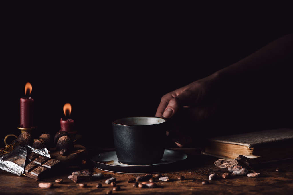 vista parziale della donna che tiene una tazza di caffè al tavolo di legno con cioccolato, tartufo, chicchi di caffè, candele e libro su sfondo nero
  - Foto, immagini