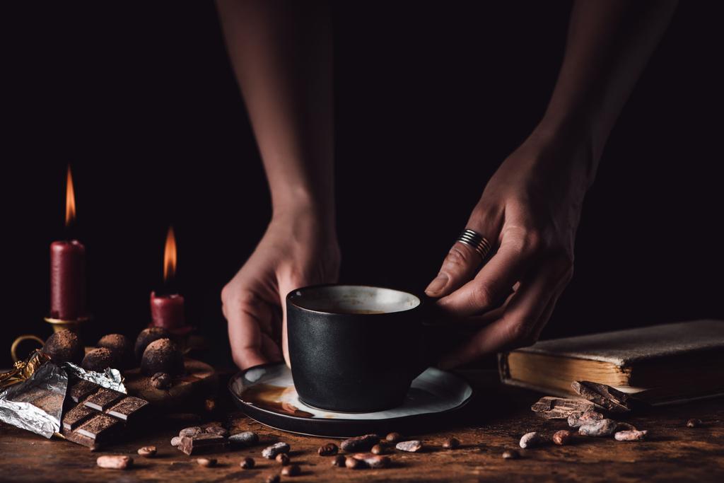 Schnappschuss einer Frau mit einer Tasse Kaffee am Holztisch mit Schokolade, Trüffeln, Kaffeekörnern, Kerzen und Buch auf schwarzem Hintergrund  - Foto, Bild
