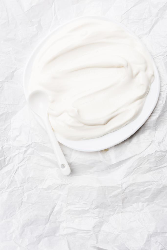 vista superior de la crema agria en el plato con cuchara sobre papel blanco arrugado
 - Foto, imagen