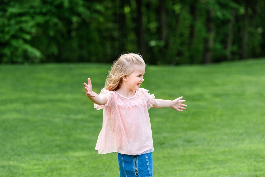 χαριτωμένο ευτυχισμένο παιδάκι με ανοιχτές αγκάλες ψάχνει μακριά στο πάρκο - Φωτογραφία, εικόνα