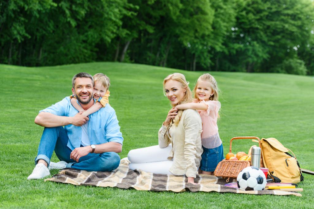 šťastná rodina s dvěma dětmi se usmívá na fotoaparátu zatímco sedí spolu na přehoz na pikniku   - Fotografie, Obrázek