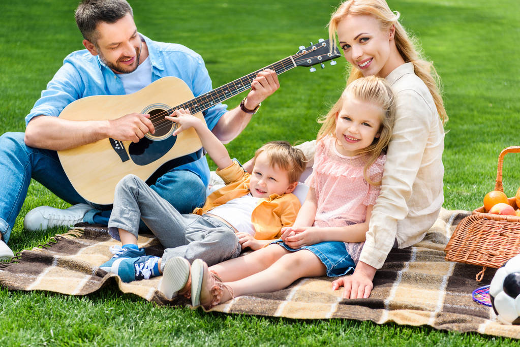 ギターを弾くとピクニックで格子縞に一緒に座っている幸せな家族  - 写真・画像