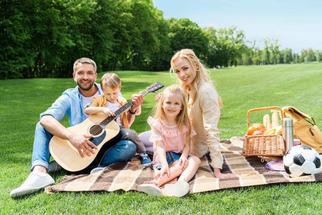 ευτυχισμένη οικογένεια με κιθάρα χαμογελώντας στην κάμερα ενώ κάθονται μαζί σε καρό σε πικ-νικ - Φωτογραφία, εικόνα