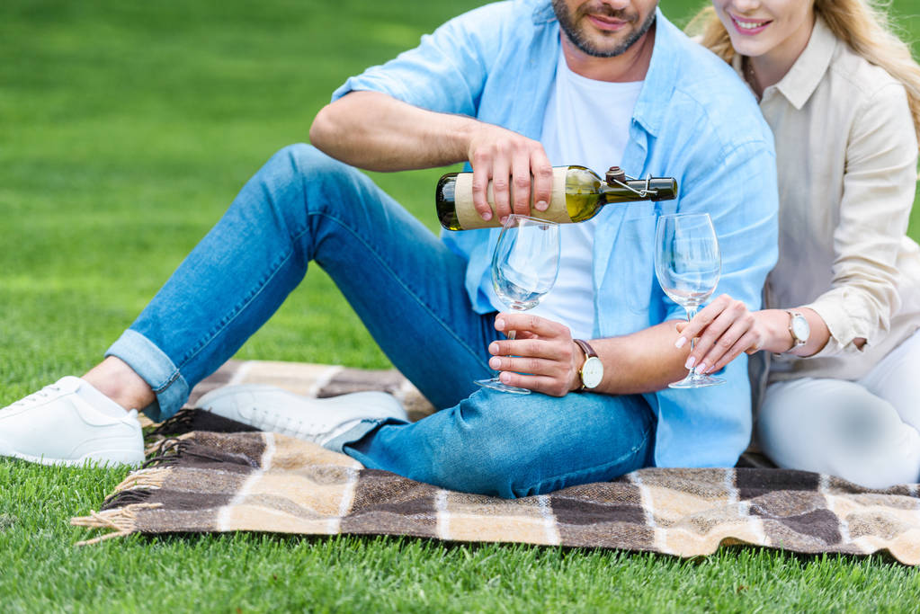 Schnappschuss von Paar, das Wein einschenkt, während es beim Picknick auf Plaid sitzt - Foto, Bild