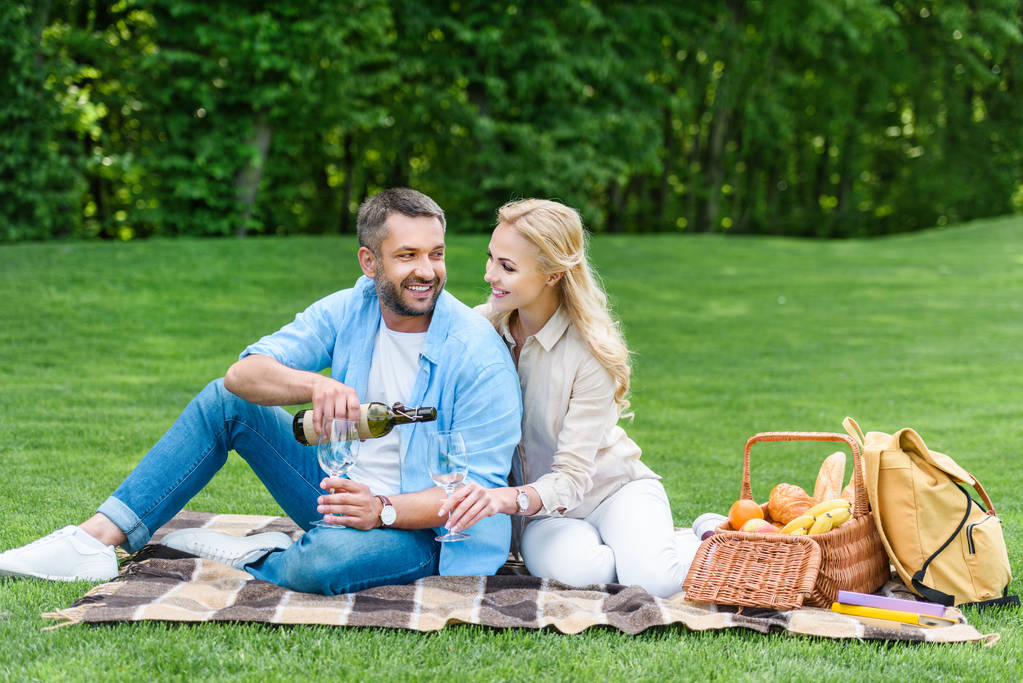 glückliches Paar trinkt Wein, während es beim Picknick zusammen auf Plaid sitzt - Foto, Bild
