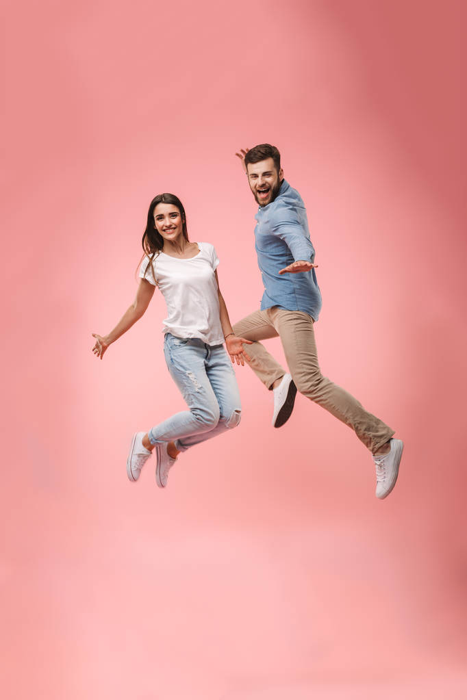 ピンクの背景に分離されたジャンプしながら手を繋いでいる陽気な若いカップルの完全な長さの肖像画 - 写真・画像