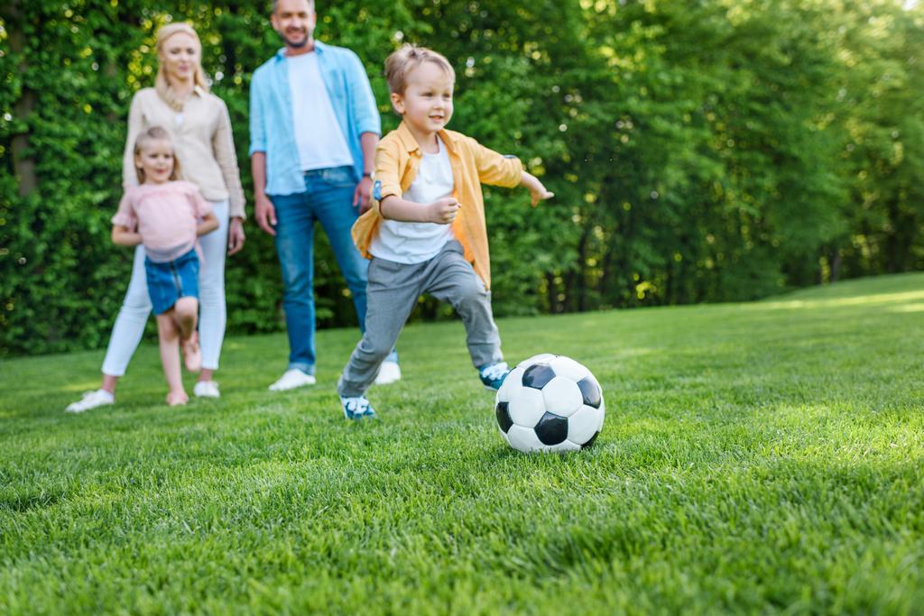 сім'я дивиться на маленького хлопчика, який грає з футбольним м'ячем у парку
 - Фото, зображення