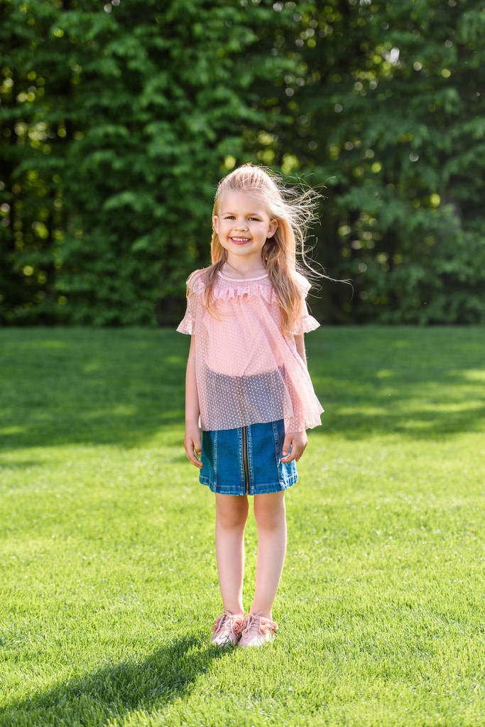 full length view of adorable little child standing on grass and smiling at camera in park - Photo, Image