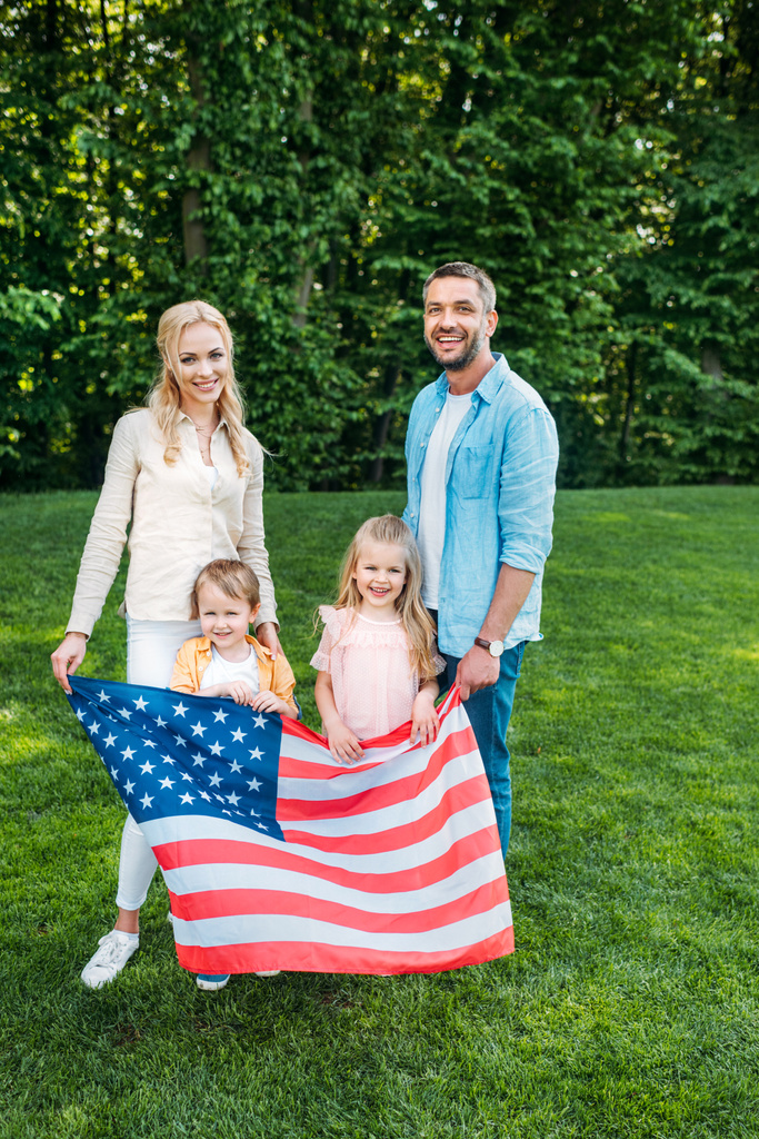 família feliz com duas crianças segurando bandeira americana e sorrindo para a câmera no parque
   - Foto, Imagem