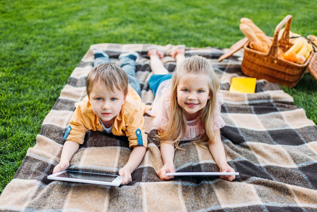 lindos niños pequeños usando tabletas digitales y sonriendo a la cámara mientras yacen en el picnic
 - Foto, Imagen