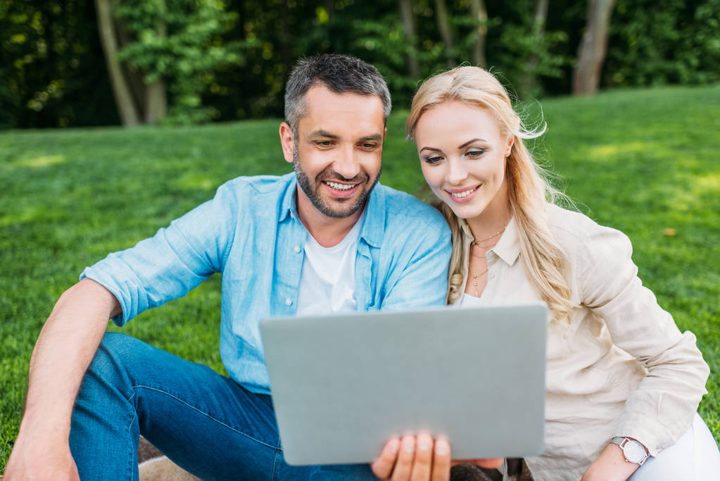 happy young couple using laptop while sitting together in park - Photo, Image