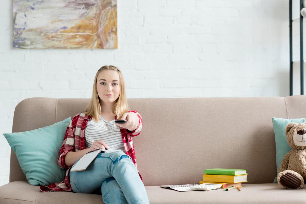 Улыбающаяся студентка с пультом дистанционного управления смотрит телевизор, сидя на диване с книгой и записками
 - Фото, изображение