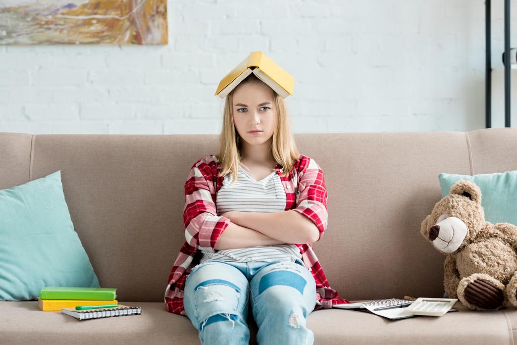 triste adolescente estudiante chica con libro en la cabeza sentado en sofá en casa y mirando a la cámara
 - Foto, Imagen