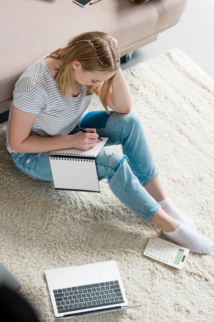 високий кут зору концентрованої дівчини-підлітка робить домашнє завдання, сидячи на підлозі
 - Фото, зображення