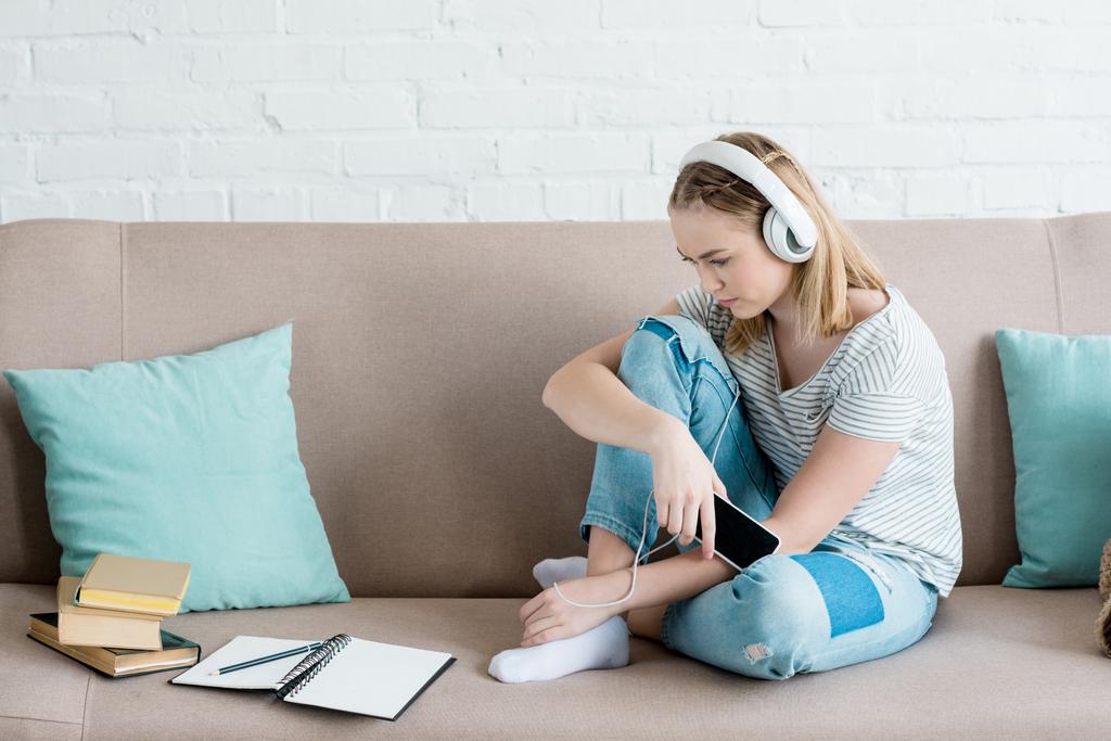 sad teen girl sitting on couch and listening music with headphones - Photo, Image