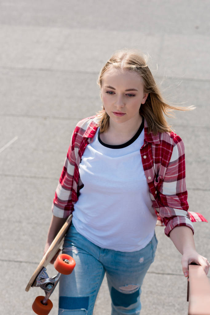 beautiful teen girl in red plaid shirt walking with skateboard in hand - Photo, Image