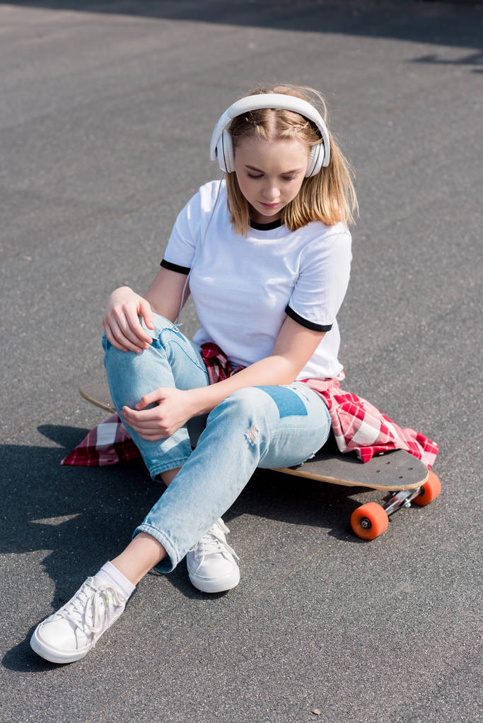 stylish teen girl listening music in headphones while sitting on skateboard - Photo, Image