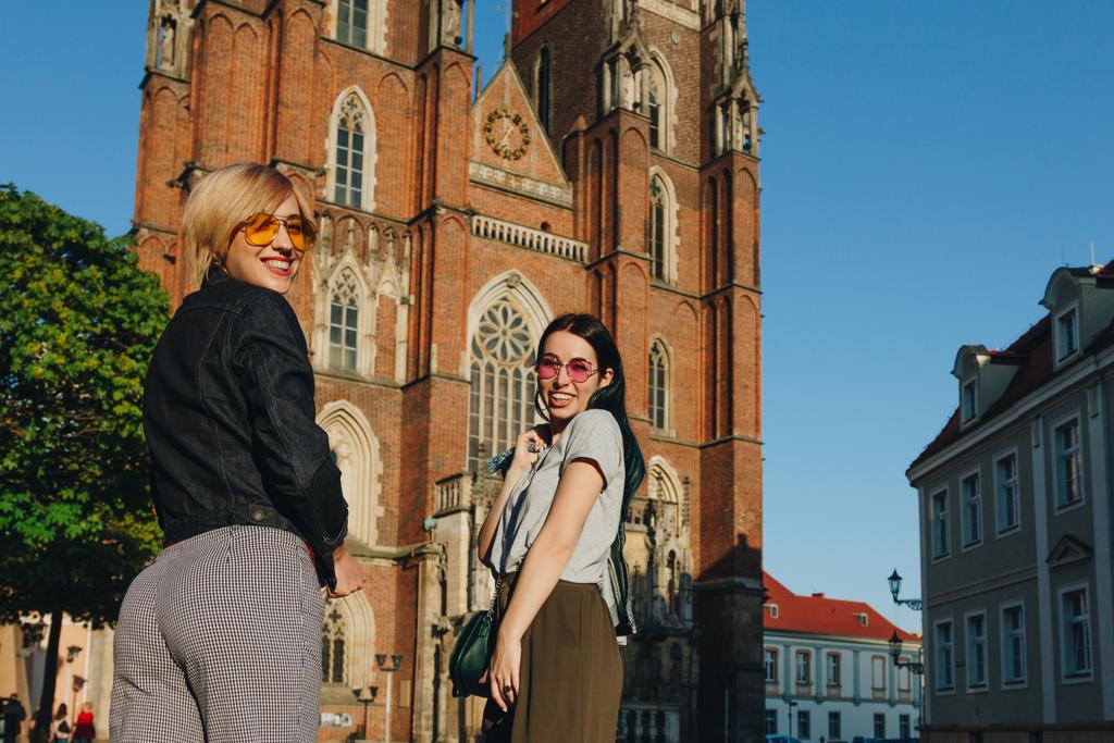 twee aantrekkelijke glimlachende vrouwen permanent voor wroclaw kathedraal en camera te kijken - Foto, afbeelding