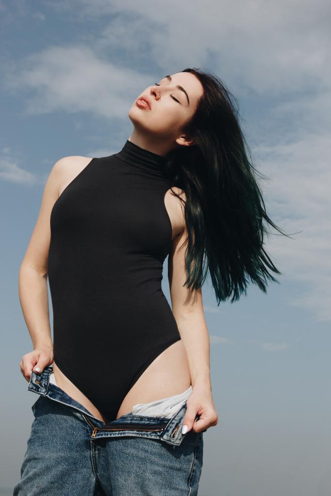 чуттєва молода жінка в чорному комбінезоні бере джинси перед хмарним небом
 - Фото, зображення