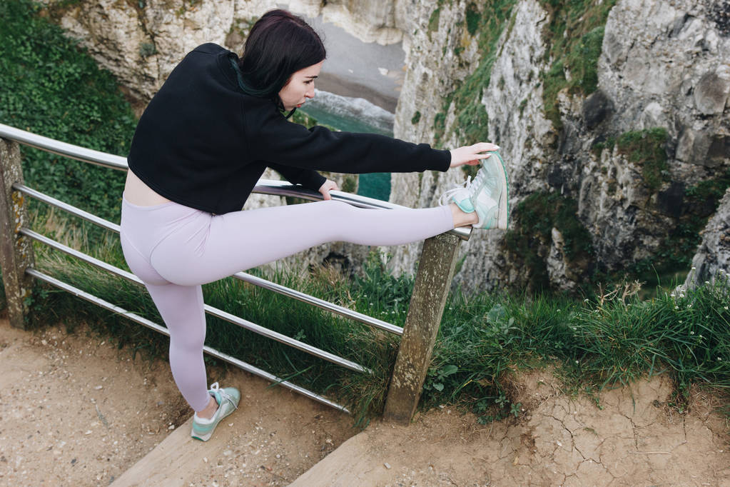 όμορφη αθλήτρια στο κομψό sportswear τεντώνοντας τα πόδια σε κάγκελα, Etretat, Γαλλία  - Φωτογραφία, εικόνα