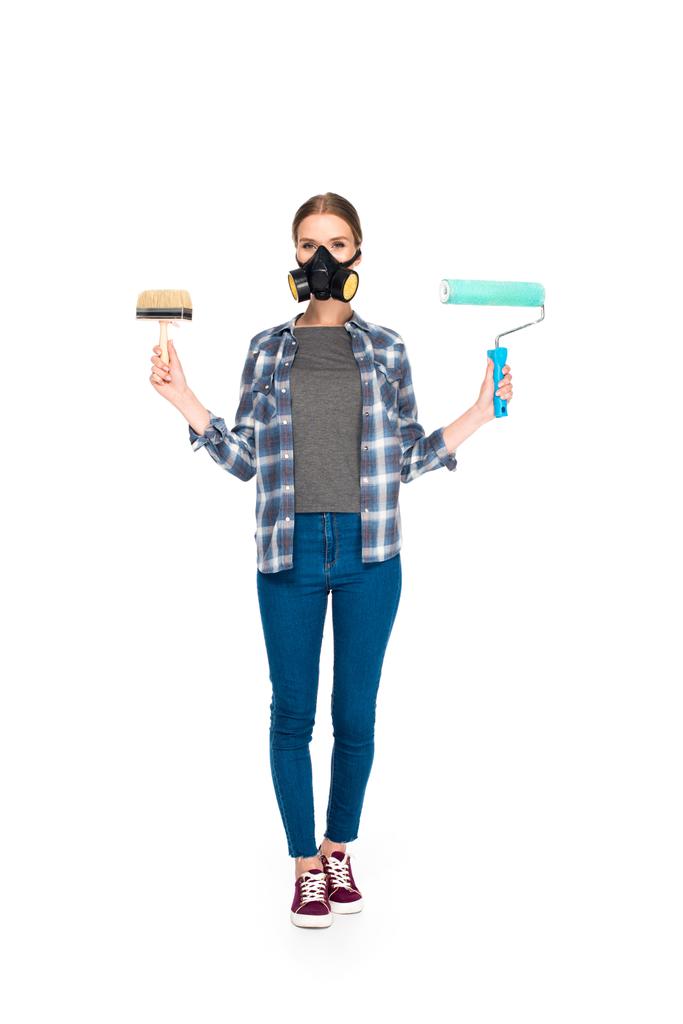 Genç kadın holding boya fırça ve boya silindiri beyaz arka plan üzerinde izole gaz maskesi  - Fotoğraf, Görsel
