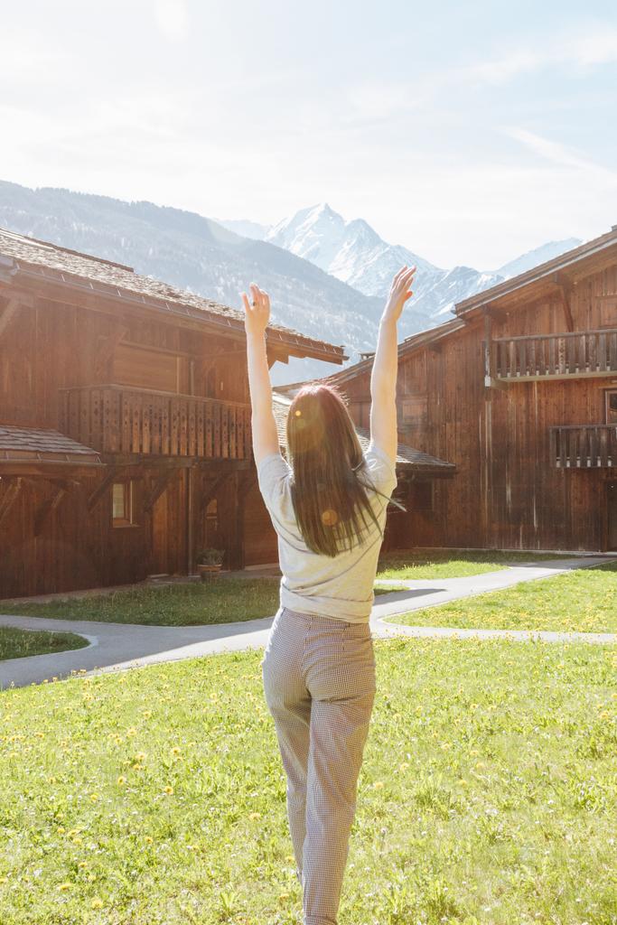 back view of beautiful young woman raising hands while standing between wooden houses in mountain village, mont blanc, alps - Photo, Image