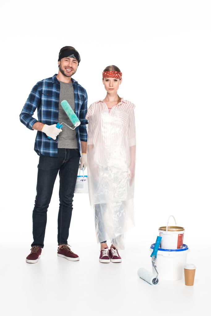 young smiling man with paint roller standing near girlfriend in polyethylene cover and painting tools with paper cup of coffee on floor isolated on white background  - Photo, Image