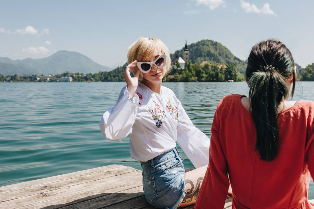 a szép lány a napszemüveg látszó-on fényképezőgép ülve barátnőjével fa mólón-a hegyi tó, bled, Szlovénia - Fotó, kép