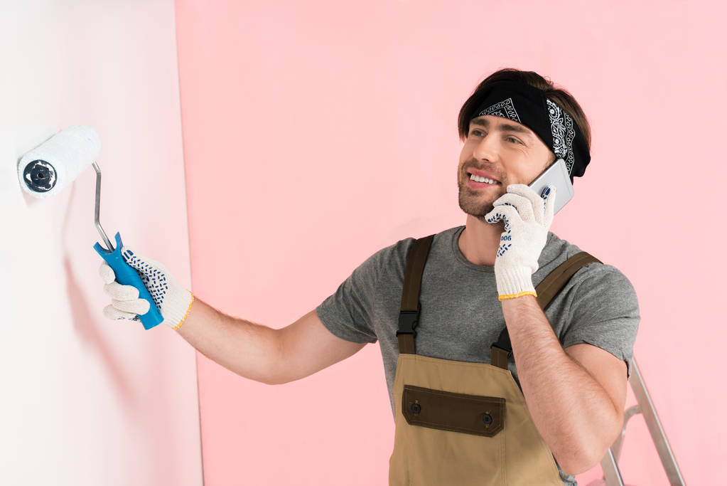 smiling man in protective gloves and working overall talking on smartphone and painting wall by paint roller  - Photo, Image