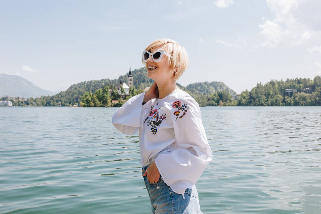 schöne lächelnde junge Frau mit Sonnenbrille, die in der Nähe des majestätischen Bergsees steht, blutend, Slowenien - Foto, Bild