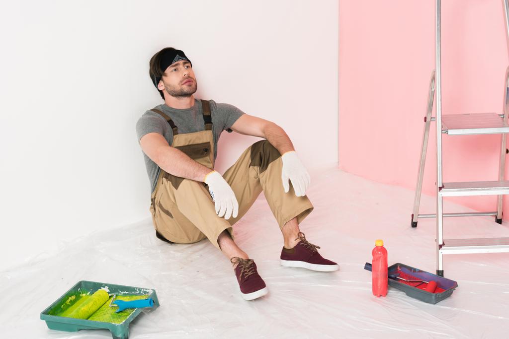 homme en bandeau et travaillant globalement reposant sur le sol près des plateaux à rouleaux avec rouleaux de peinture, bouteille et échelle
  - Photo, image