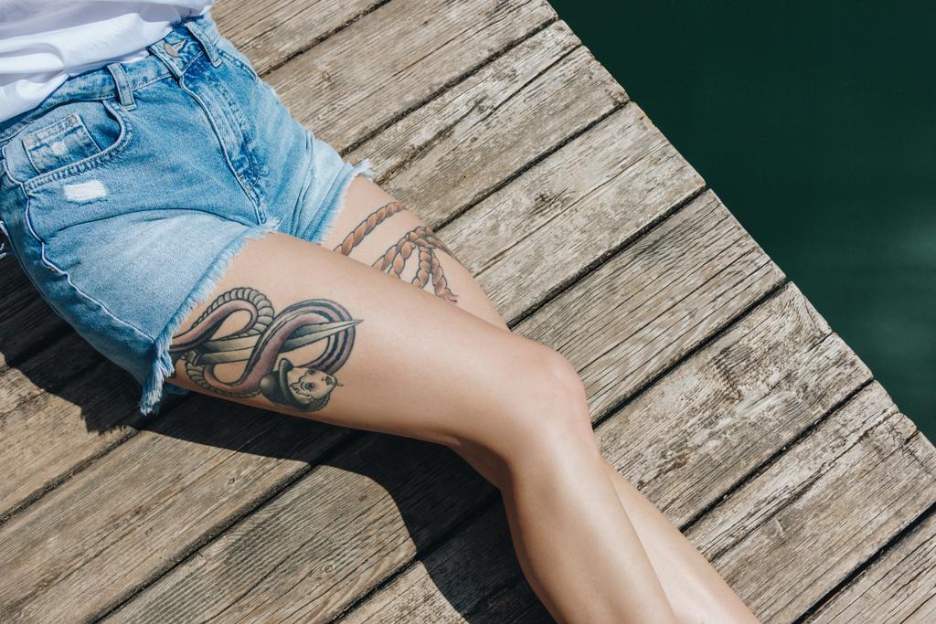 vista dall'alto parziale di una giovane donna in pantaloncini di jeans appoggiata su un molo di legno vicino all'acqua
 - Foto, immagini
