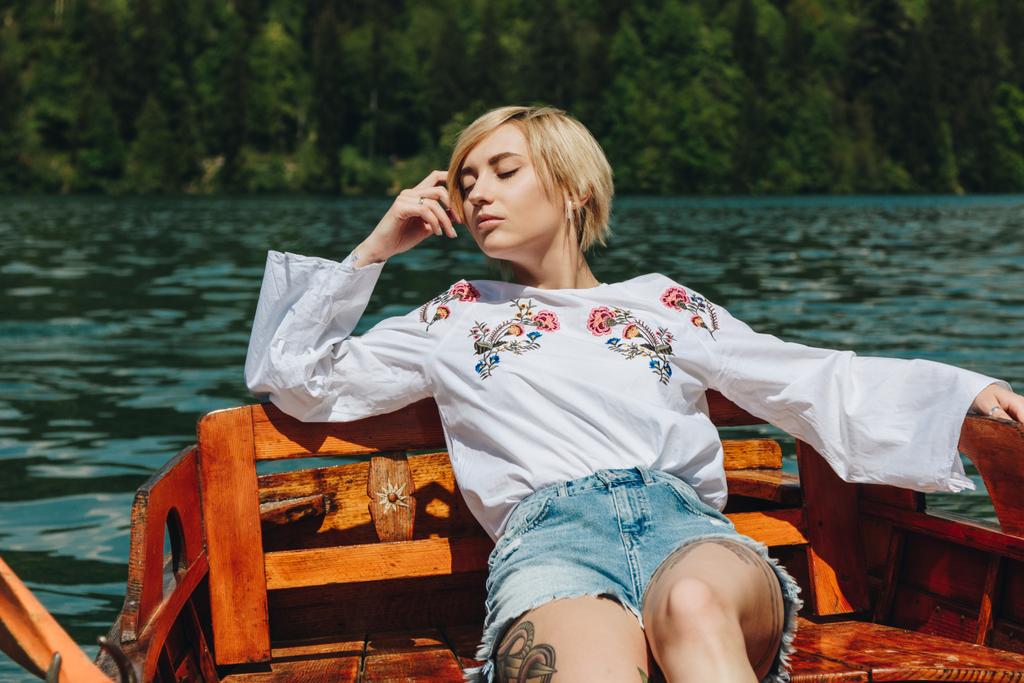 piękna młoda kobieta z zamkniętymi oczami siedząc w drewnianej łodzi na jeziorze - Zdjęcie, obraz