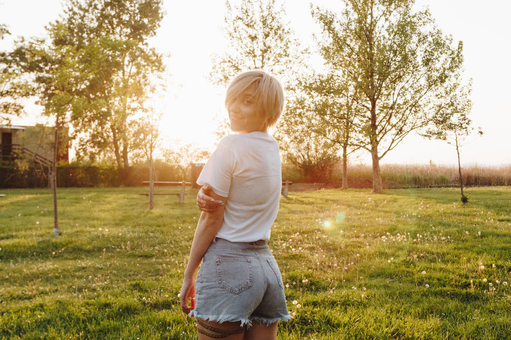 belle jeune femme en t-shirt et short en denim regardant la caméra tout en se tenant debout sur la pelouse verte au coucher du soleil
 - Photo, image