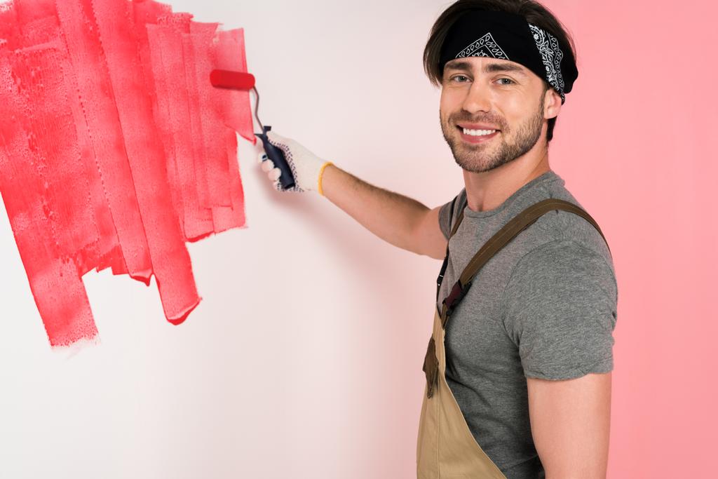 Lächelnder Mann im Arbeitsanzug und Stirnband, Wand in Rot mit Farbwalze bemalt  - Foto, Bild