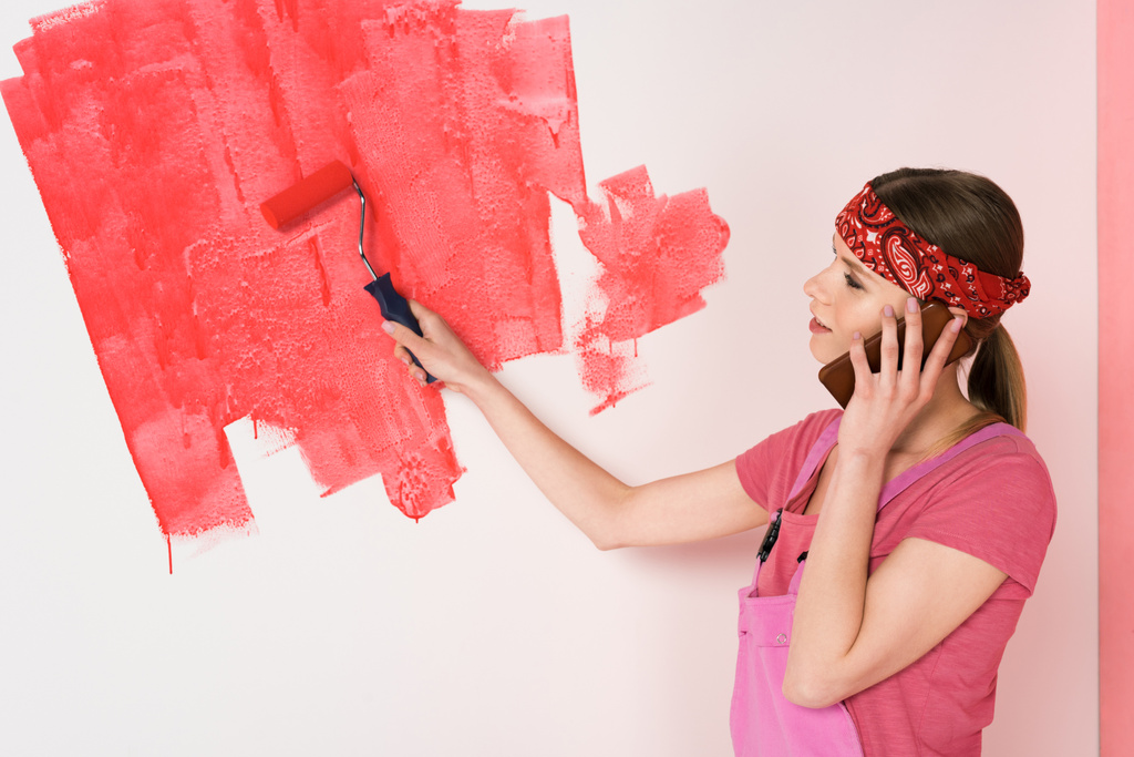 visão lateral da jovem mulher na cabeça e trabalhando em geral falando no smartphone e pintura parede em vermelho por rolo de pintura
  - Foto, Imagem