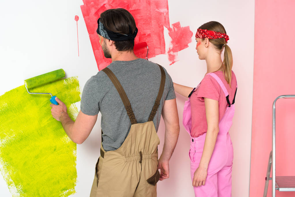 vista trasera de pareja joven en overoles de trabajo pintura pared por rodillos de pintura cerca de escalera
  - Foto, Imagen