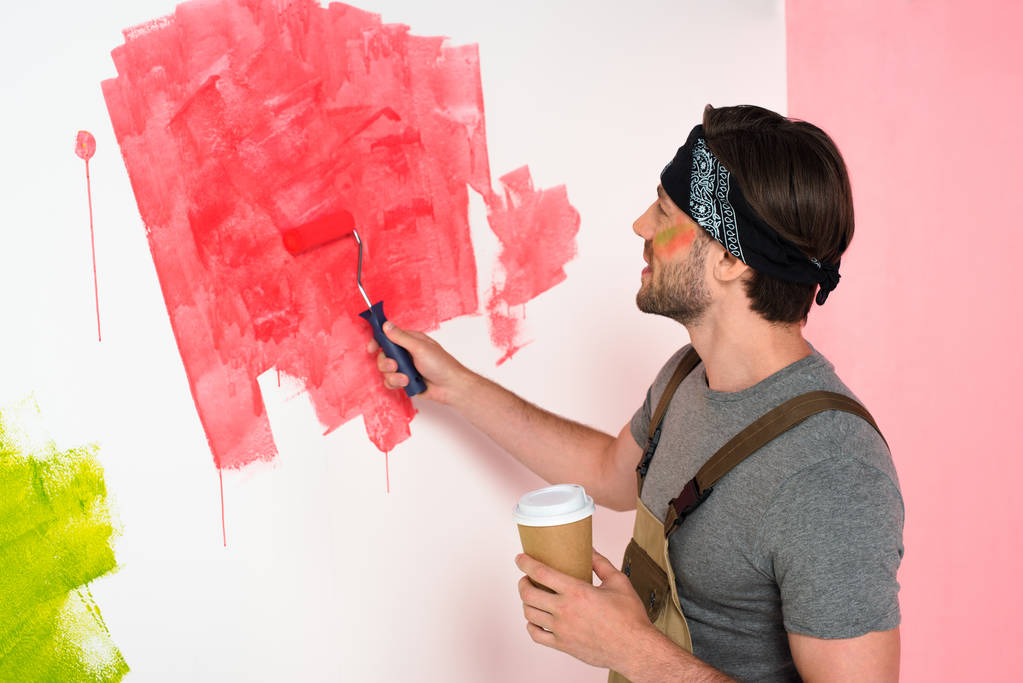 vista lateral del hombre sonriente con la pared de pintura taza de café en rojo por rodillo de pintura
 - Foto, imagen