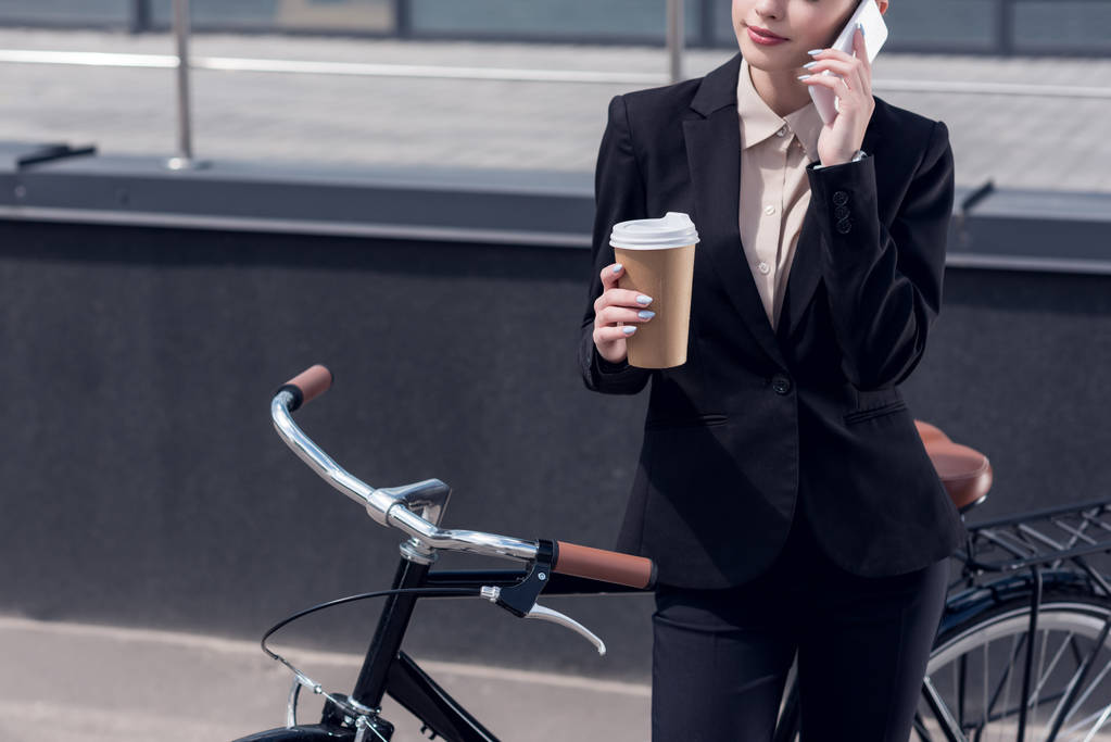 vista parcial de la mujer de negocios con café para ir a hablar en el teléfono inteligente, mientras que de pie cerca de bicicleta retro en la calle
 - Foto, imagen