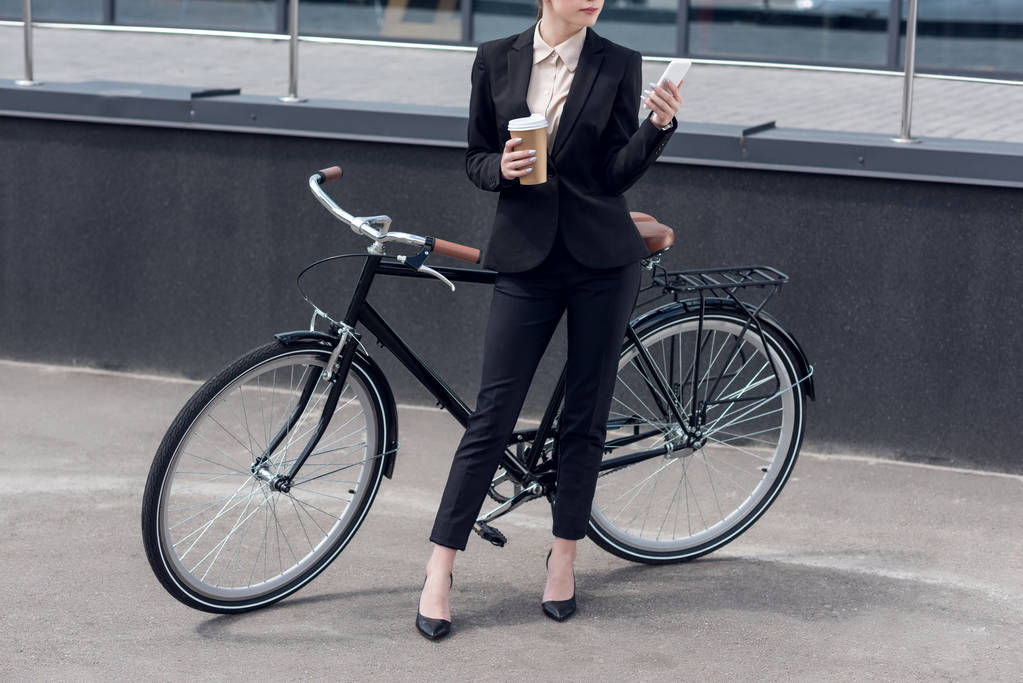 Teilbild einer Geschäftsfrau mit Coffee to go und Smartphone, die neben einem Retro-Fahrrad auf der Straße steht - Foto, Bild