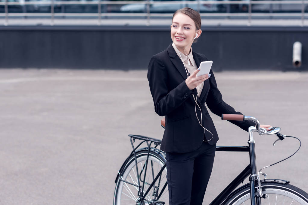 Portret van Glimlachende zakenvrouw in oortelefoons met smartphone staande in de buurt van retro fiets op straat - Foto, afbeelding