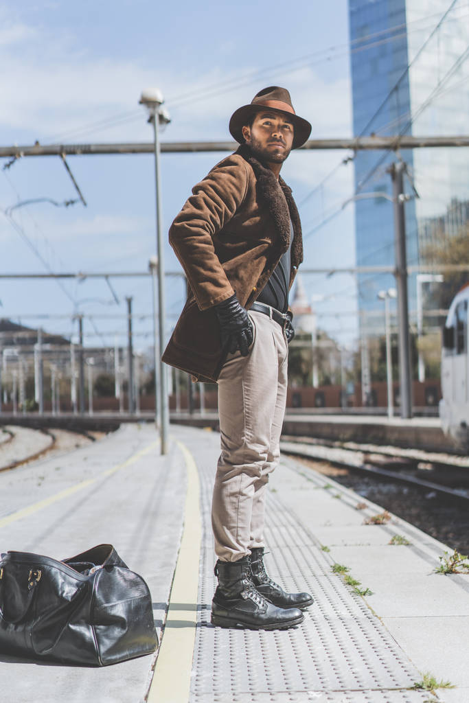 Привабливий впевнений іспаномовний чоловік у капелюсі, що має сумку для одягу за спиною, чекає потяг на платформі
. - Фото, зображення
