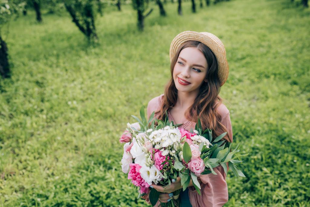 retrato de mujer joven sonriente en sombrero con ramo de flores mirando hacia el parque
 - Foto, Imagen