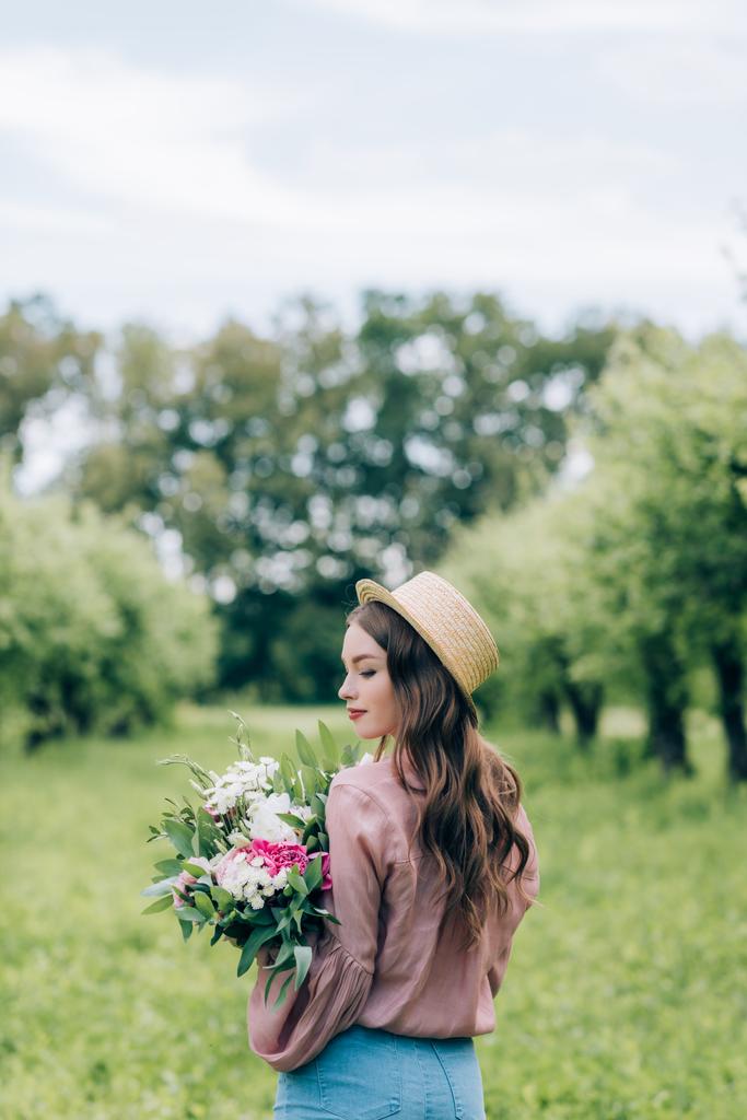Rückansicht einer jungen Frau mit Hut und Blumenstrauß in den Händen, die im Park steht - Foto, Bild