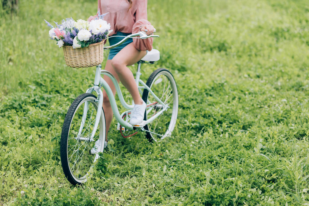 Teilansicht einer Frau auf Retro-Fahrrad mit Weidenkorb voller Blumen im Wald - Foto, Bild