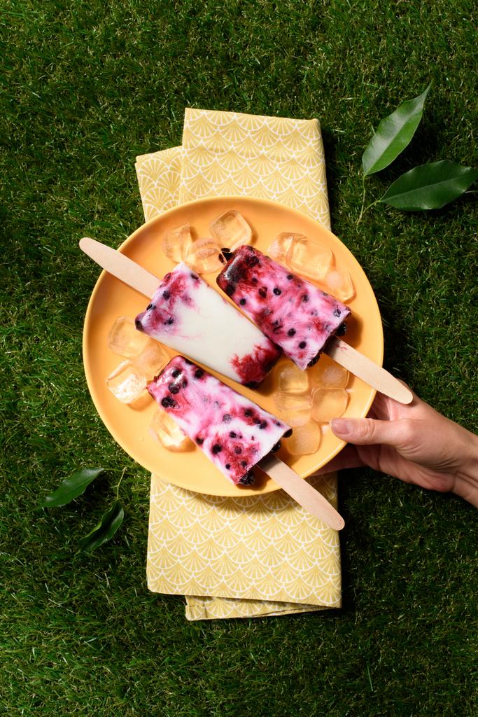 Woman holding plate with sweet fruit Popsicles on green grass background - Photo, Image