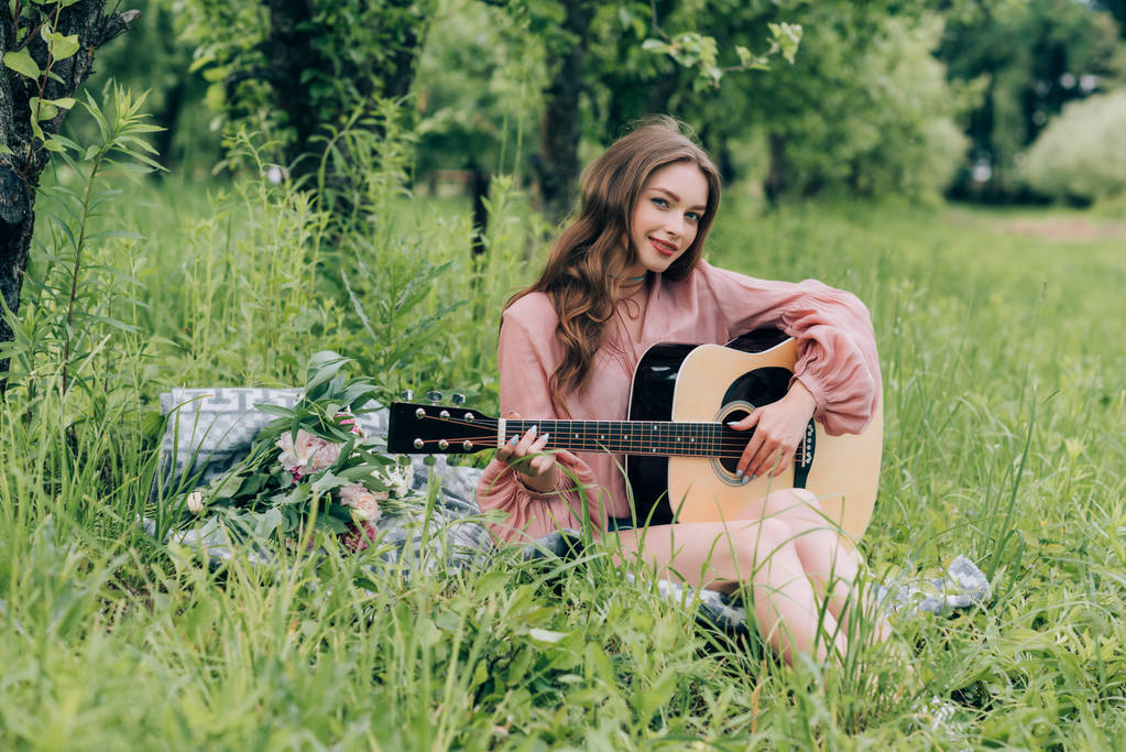 young smiling woman with acoustic guitar resting on blanket with bouquet of flowers in park - Photo, Image